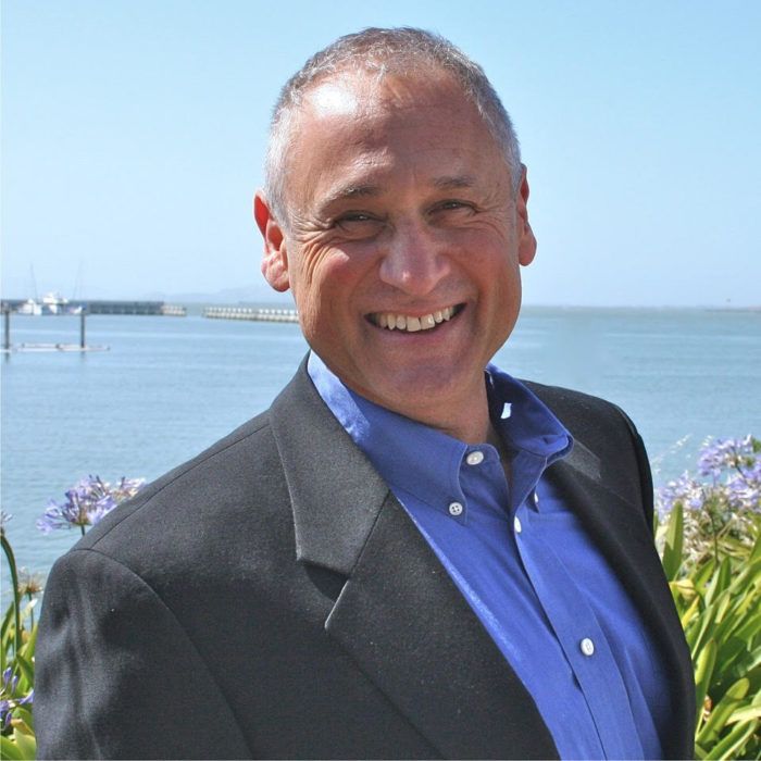 A man in a suit and blue shirt smiles in front of a body of water