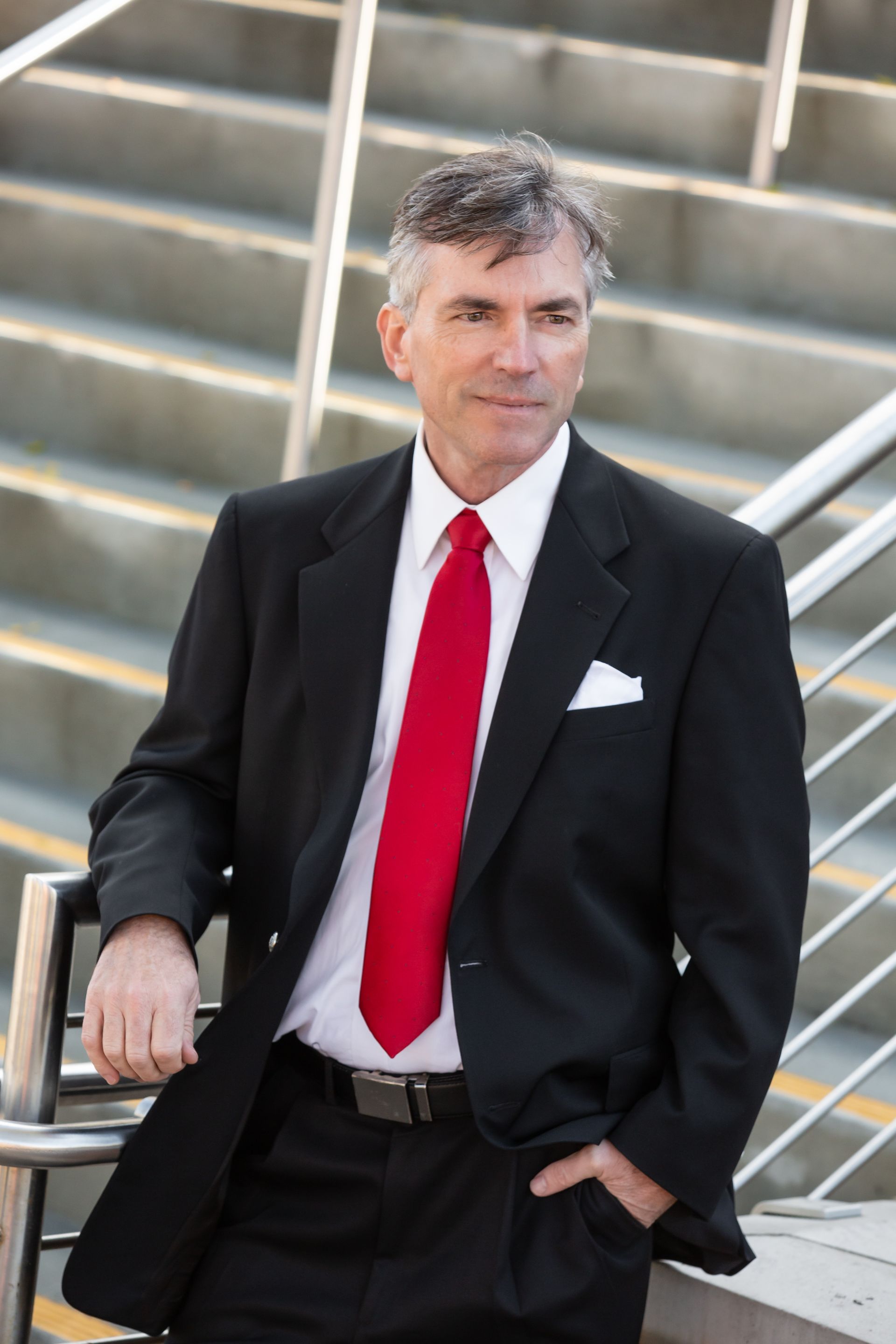 A man in a suit and red tie is leaning against a railing
