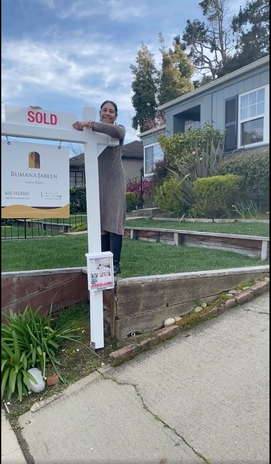 A woman is standing next to a sold sign in front of a house.