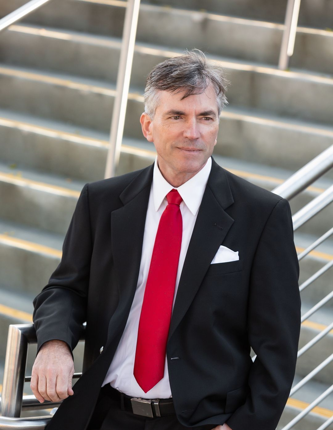 A man in a suit and red tie is leaning against a railing