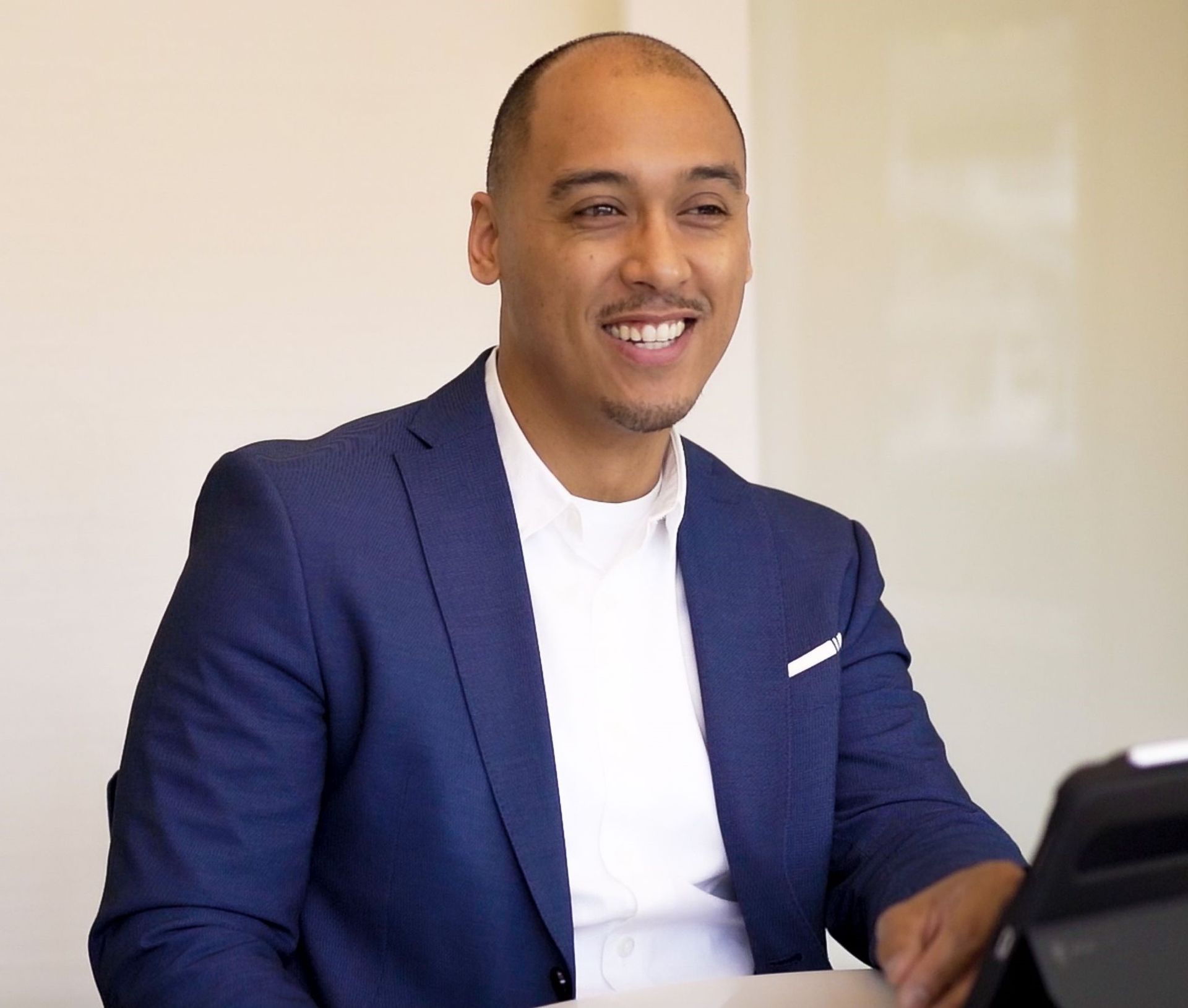 A man in a blue suit and white shirt is smiling while sitting at a desk.