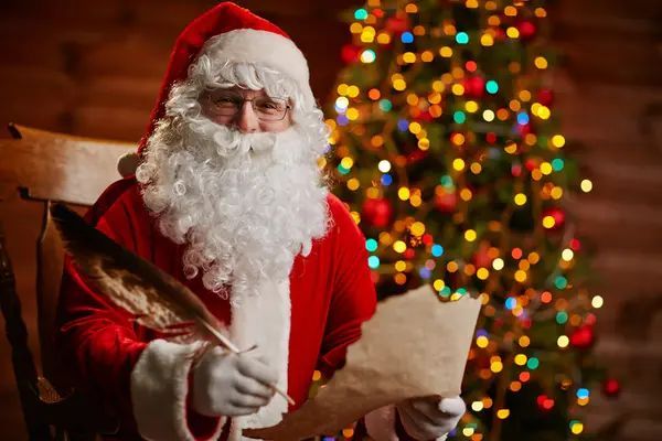 Santa claus is writing a letter to a child in front of a christmas tree.