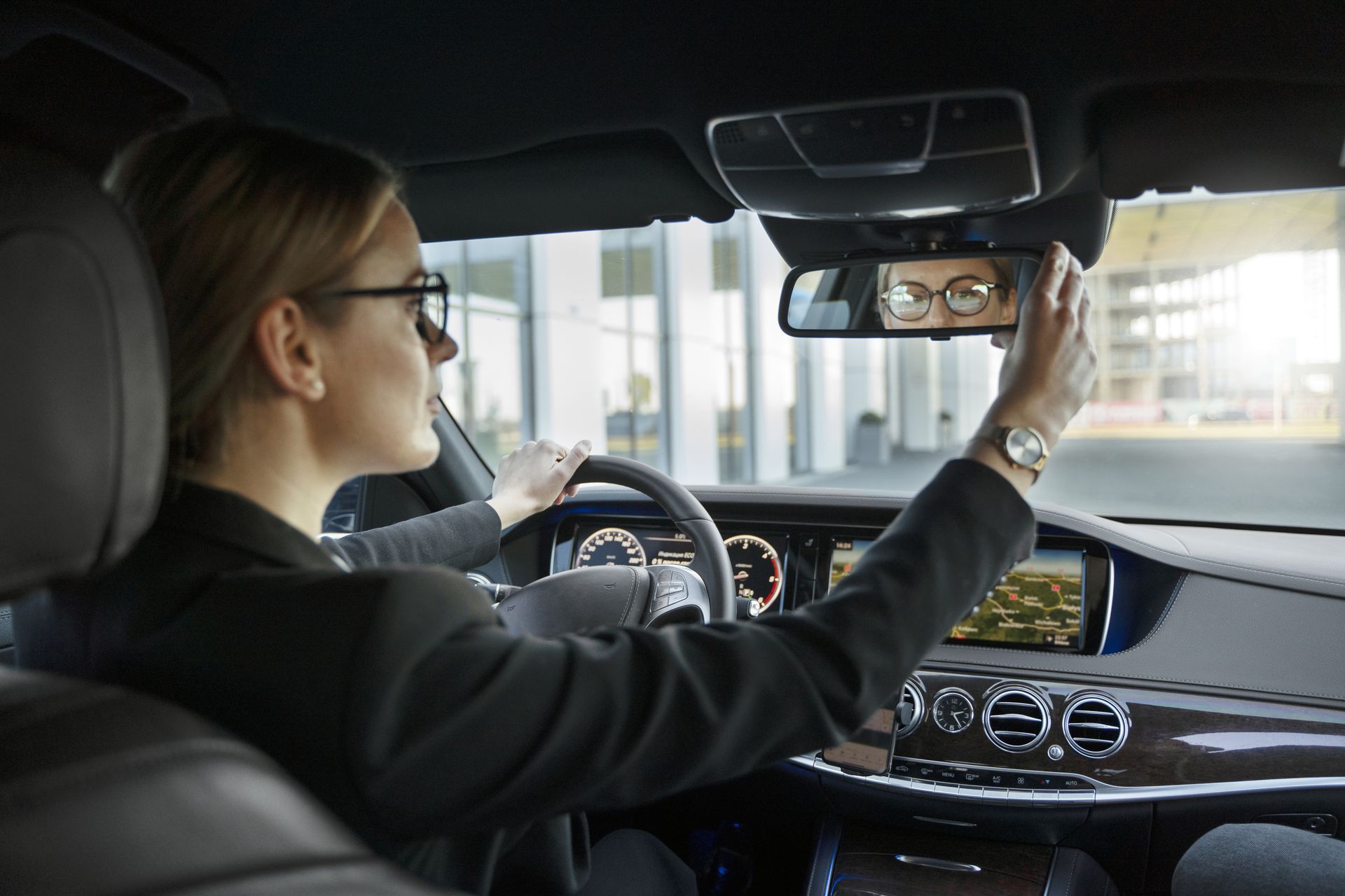Mujer con gafas ajustando el retrovisor de un coche. Se ven el salpicadero y el edificio.