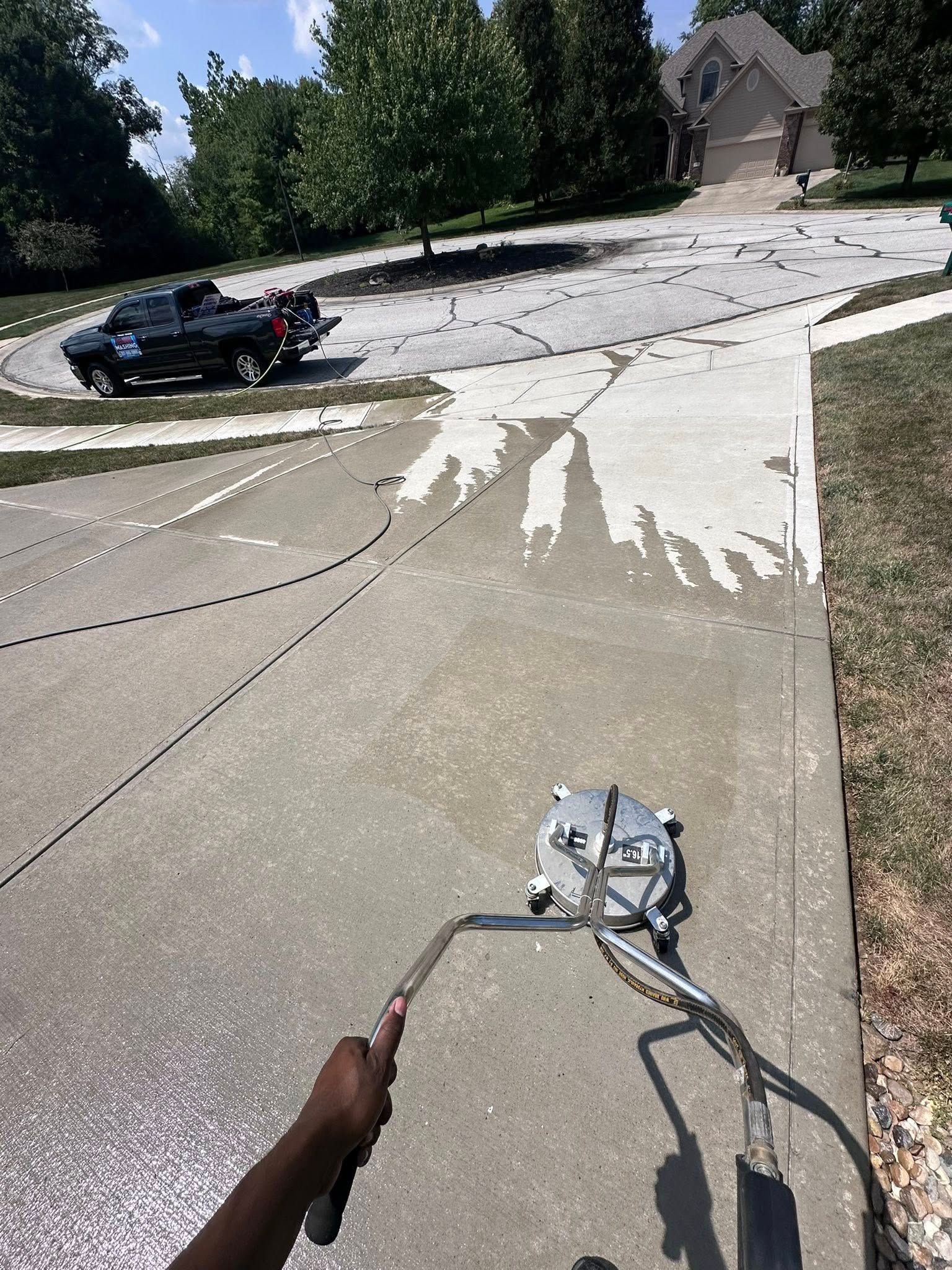 Worker painting a sunlit driveway with a spray wand, black car parked nearby, suburban house in background
