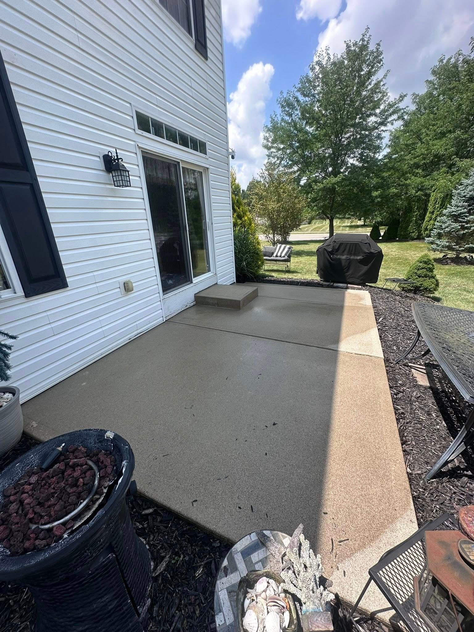Concrete patio beside white house with grill, potted plant, and landscaped yard under blue sky