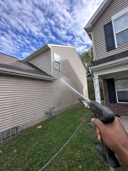 Hand spraying water at vinyl siding beside a house and garage under a blue, cloudy sky