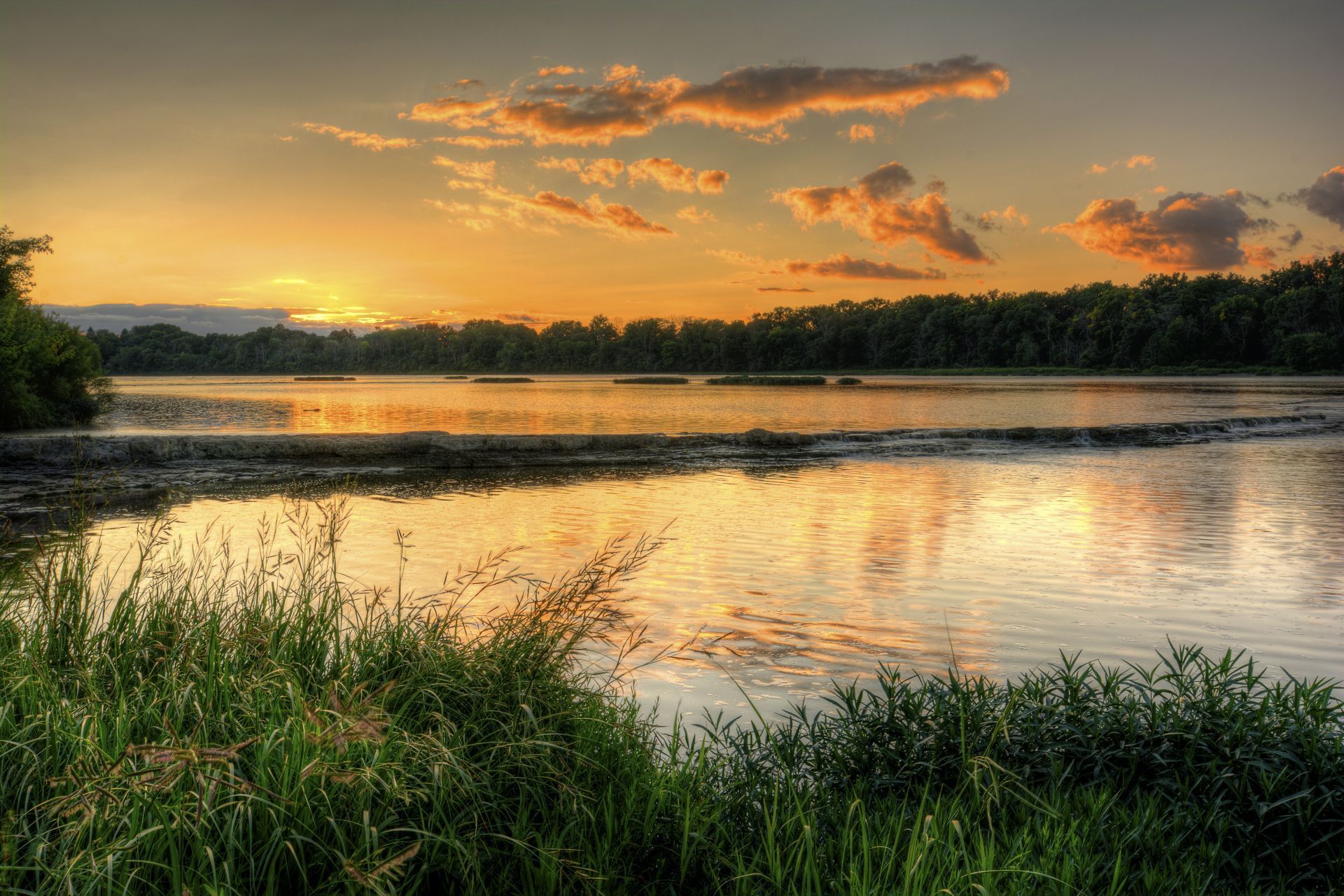 The sun is setting over a grassy field with a wooden fence in the foreground.