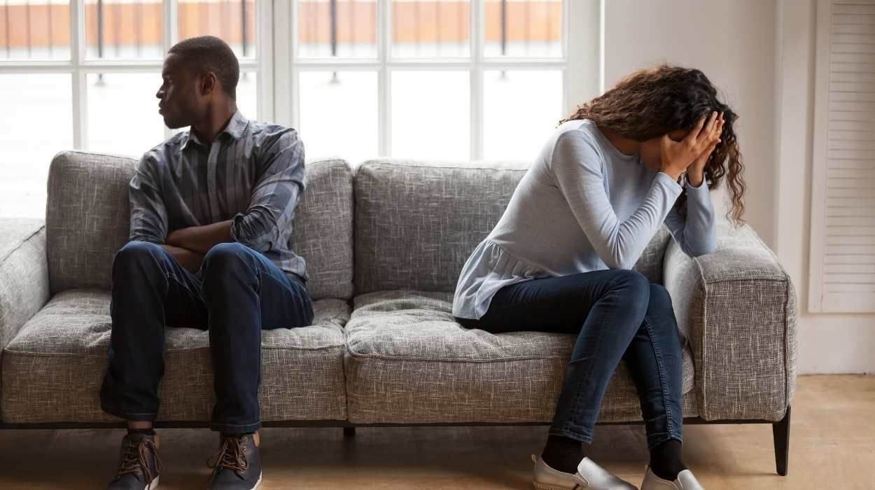 Two people sit on opposite ends of a gray sofa, facing away from each other with frustrated expressions.
