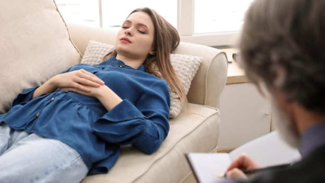 A person lies resting on a sofa while a professional sits nearby, taking notes in a notebook during a session.