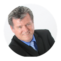 A man wearing a black suit jacket and blue dress shirt with a tie, smiling against a plain white circular background.