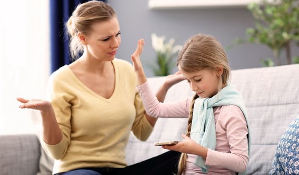 A person in a yellow sweater gestures with open hands toward a child on a sofa who is ignoring them to use a smartphone.