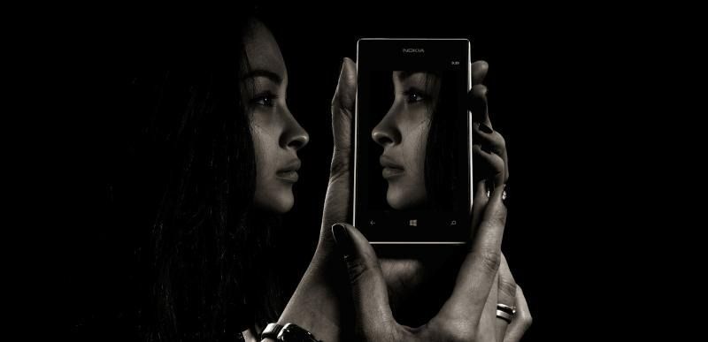 A person holds a smartphone toward their face, reflecting their profile on the screen against a dark background.