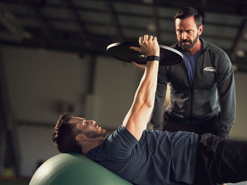 A man on a fitness ball presses a weight plate upward while being spotted by a coach in a gym setting.