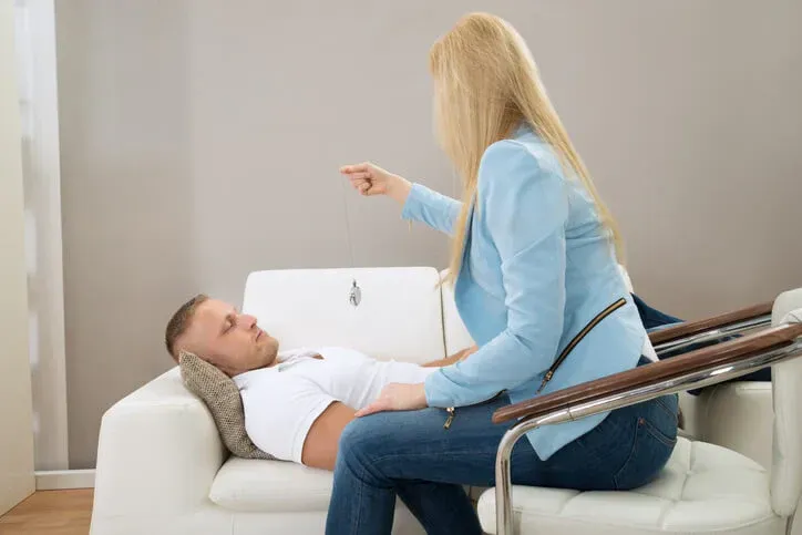 A person sits on the edge of a chair, pointing toward a person lying on a white sofa in a bright, modern room.