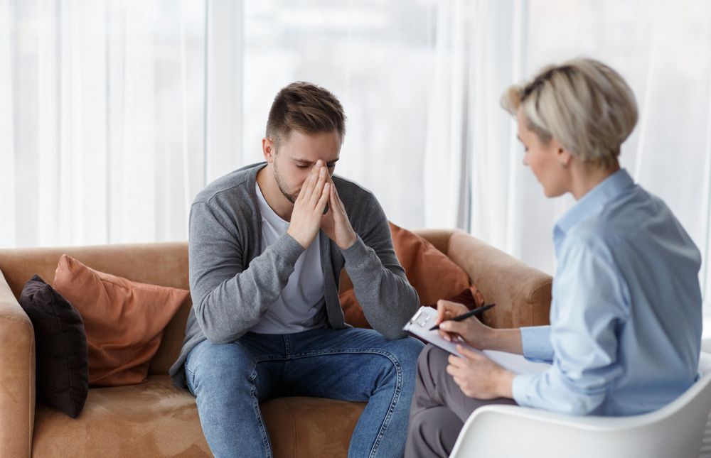 A distressed person sits on a sofa with hands to their face while a counselor takes notes during a therapy session.