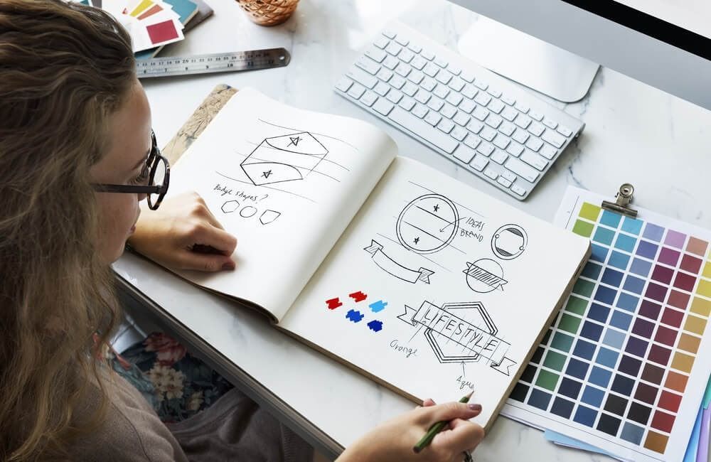 A woman is sitting at a desk drawing in a notebook.