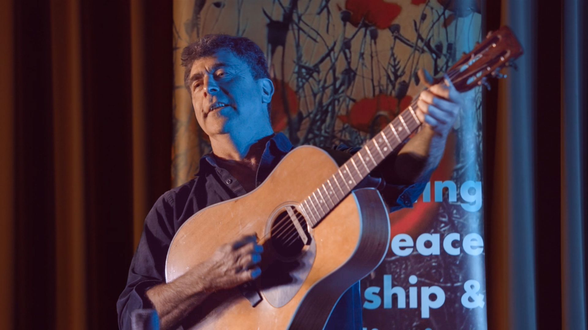 A man is playing an acoustic guitar in front of a sign that says peace ship