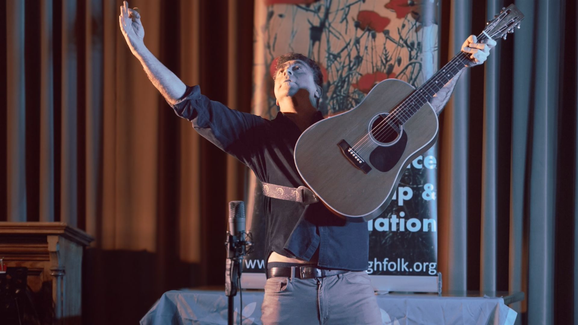 A man is holding a guitar in front of a microphone with his arms outstretched.