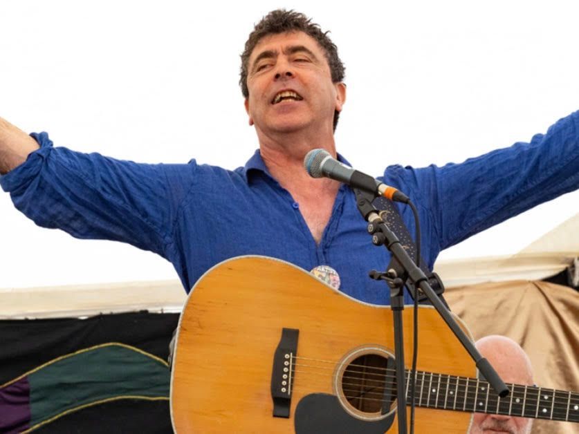 Man singing and playing acoustic guitar with arms outstretched. Blue shirt, stage microphone.