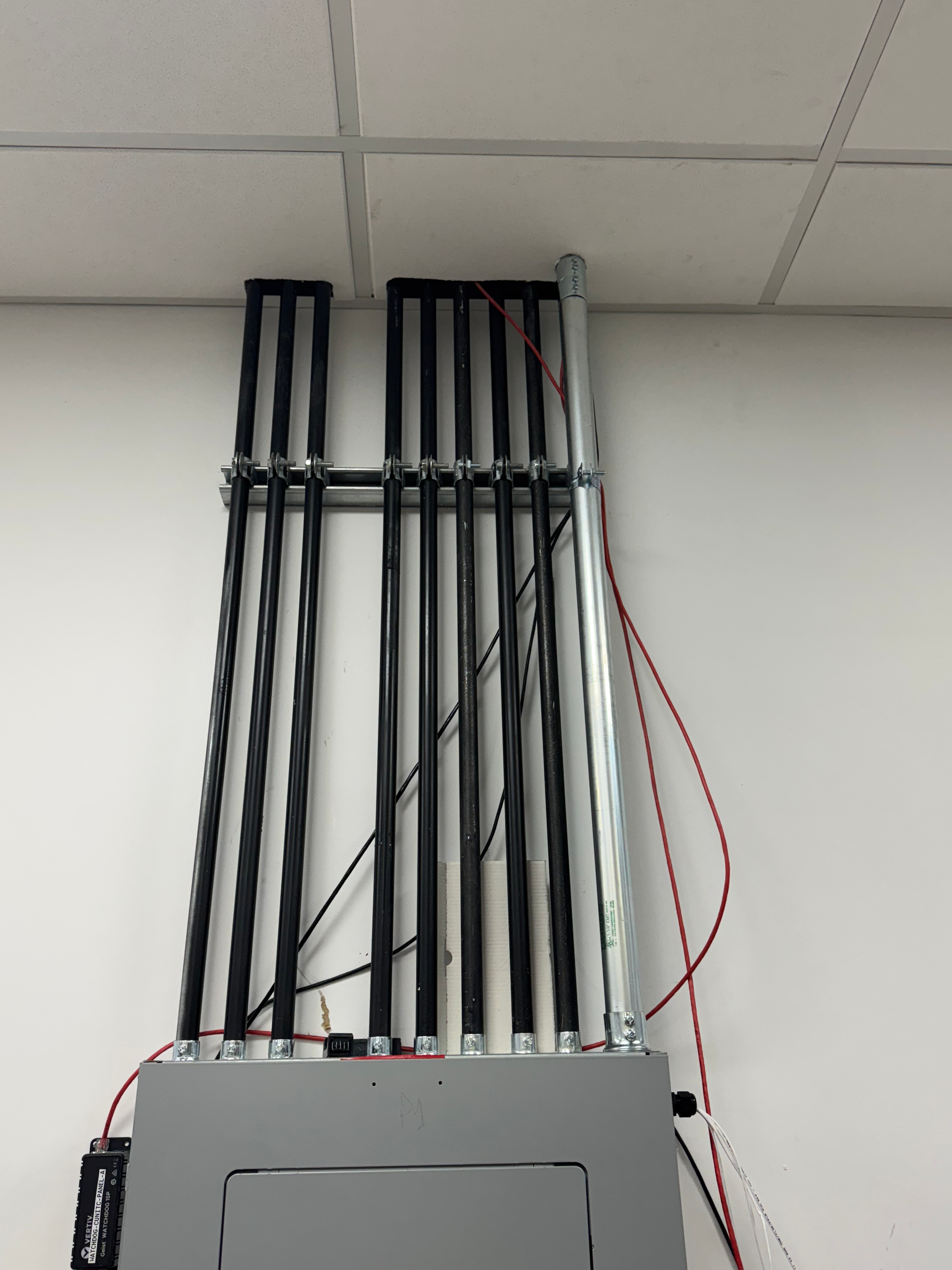 Vertical black cables and red wires running along a ceiling wall above a gray panel箱