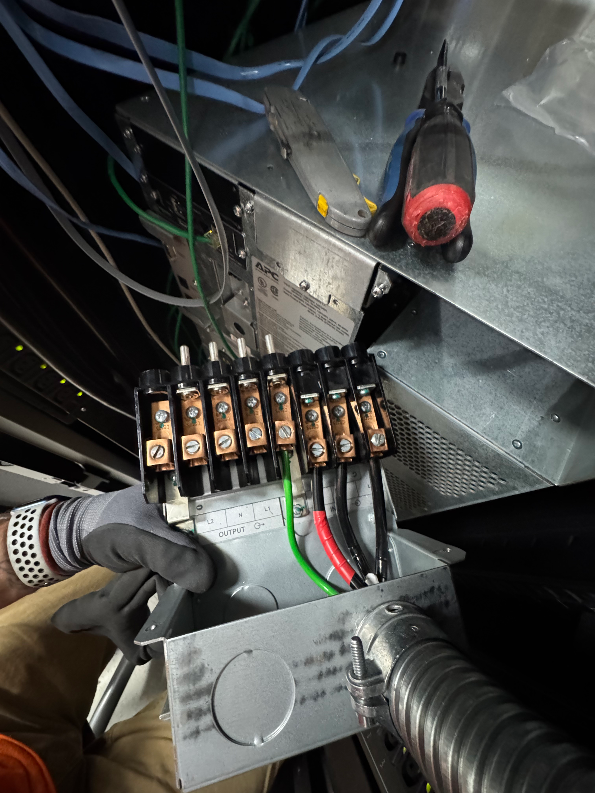 Gloved hand works on an electrical panel with wires, terminal blocks, and a red flashlight inside a metal enclosure