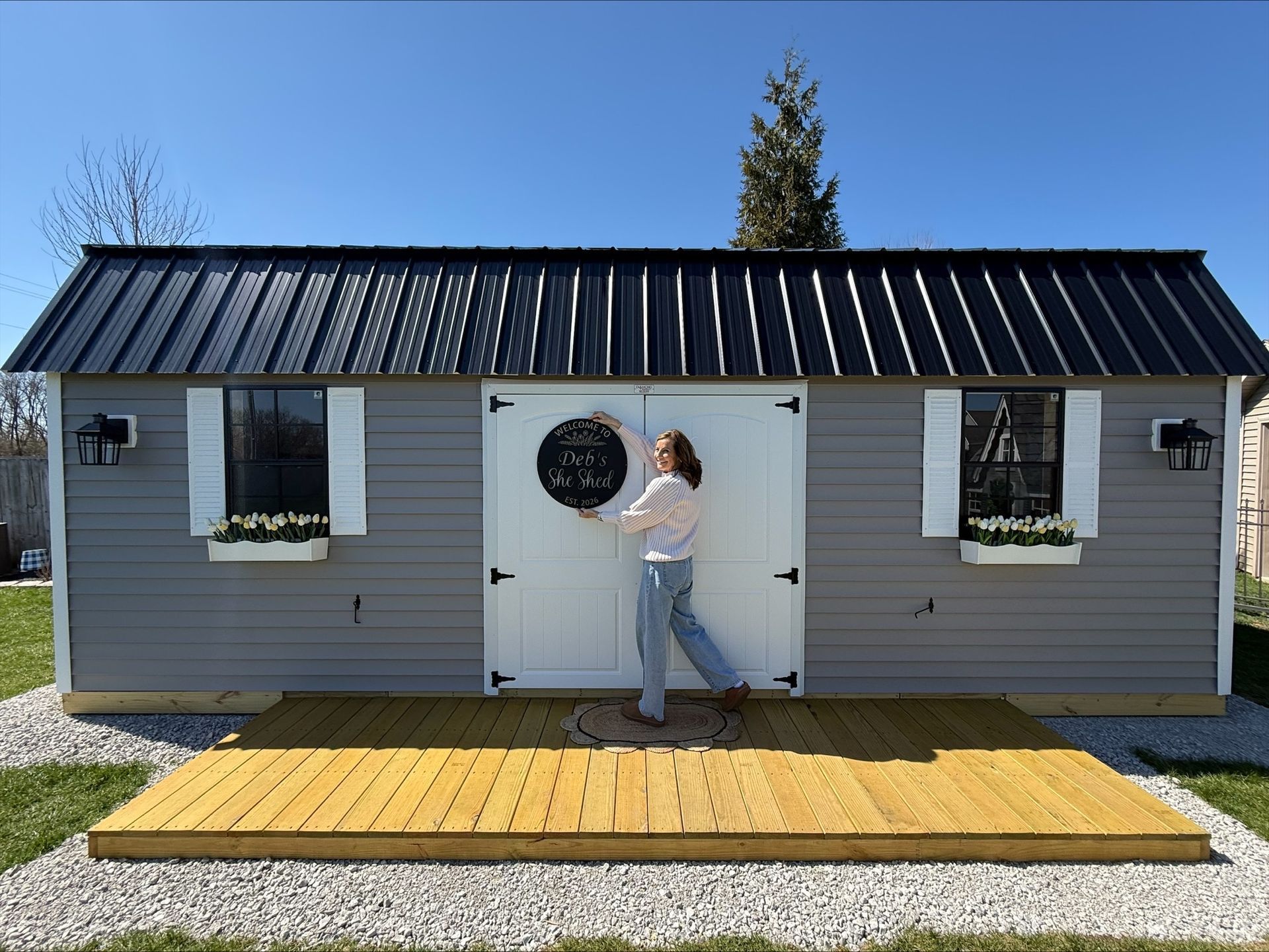 Gray shed-style tiny house with black roof and white doors, a person standing on the wooden deck outside.