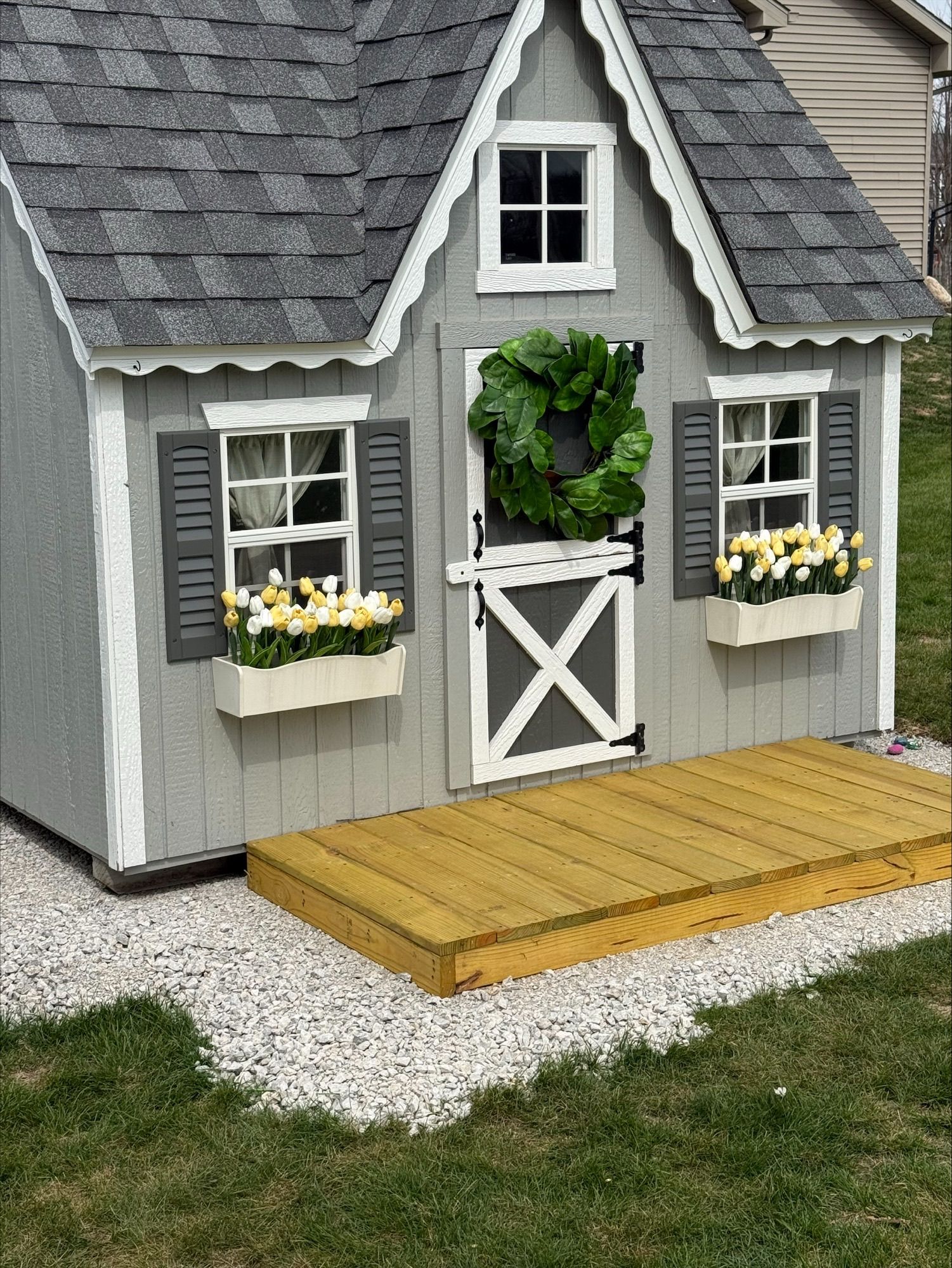 Small gray-and-white dollhouse with flower boxes and a wooden porch on gravel.