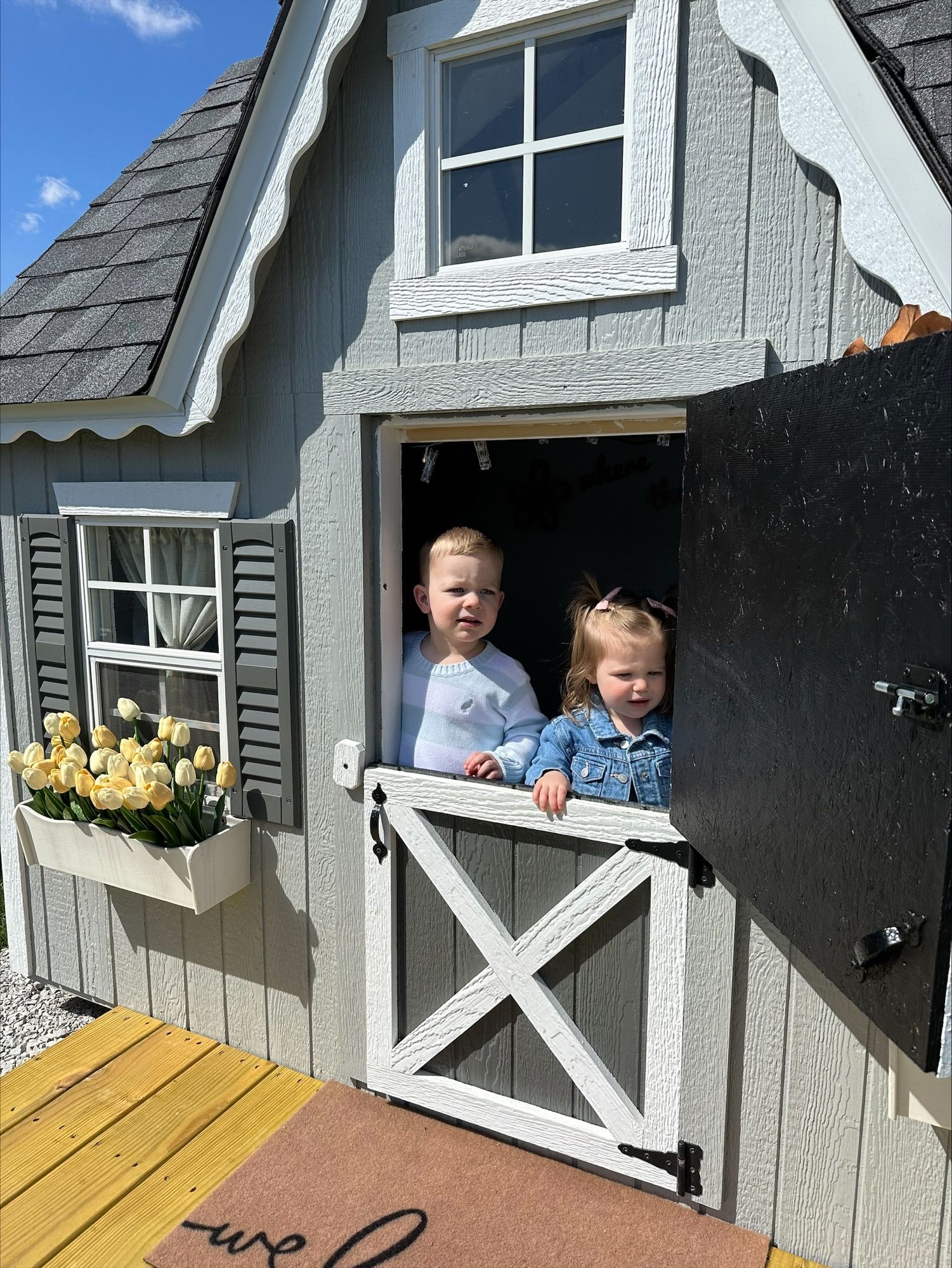 Two children leaning out of a cottage-style window with flowers in a planter and a half-open black shutter