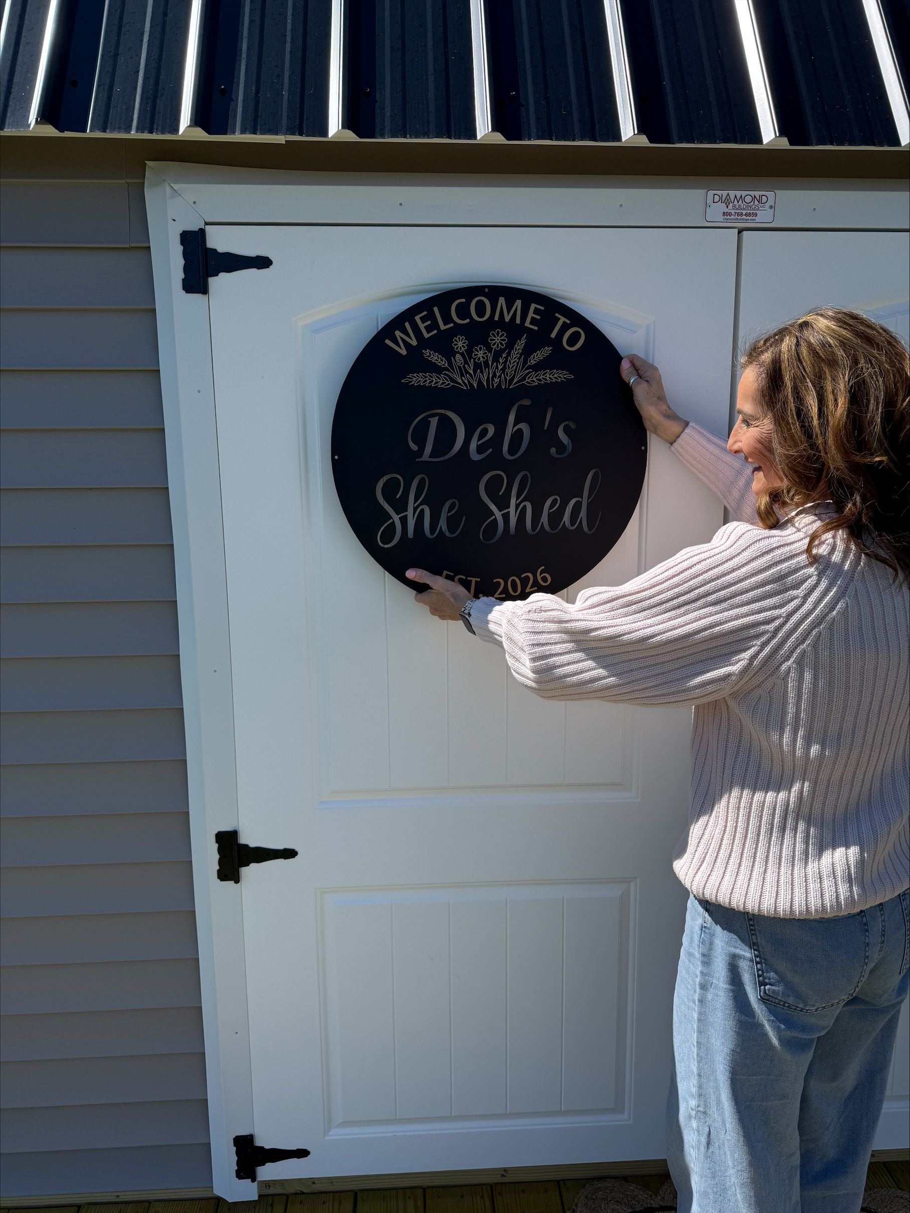 Woman opening a white shed door with a round Deb’s She Shed sign.