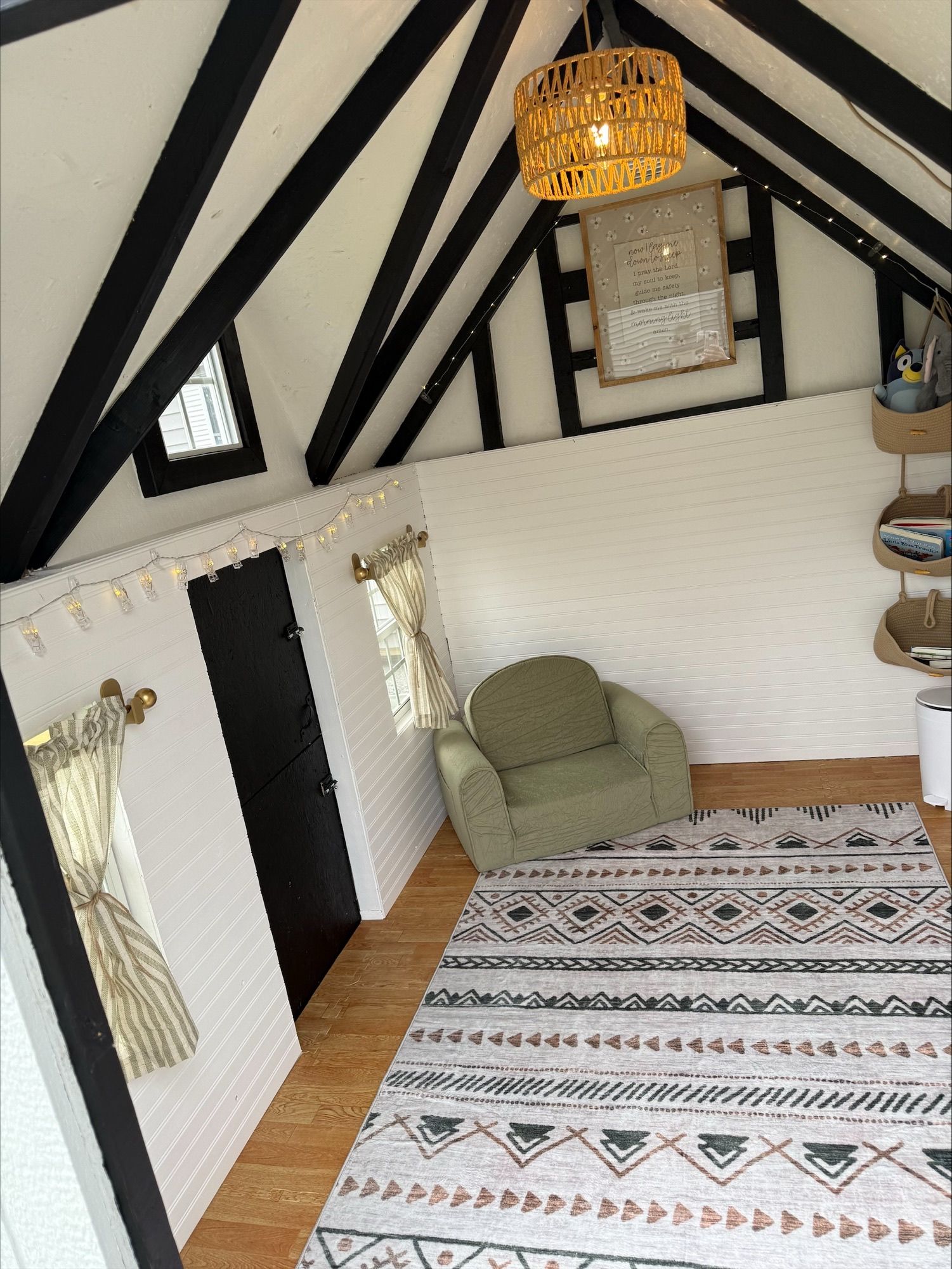 Cozy attic room with exposed black beams, green chair, patterned rug, and hanging light fixture.
