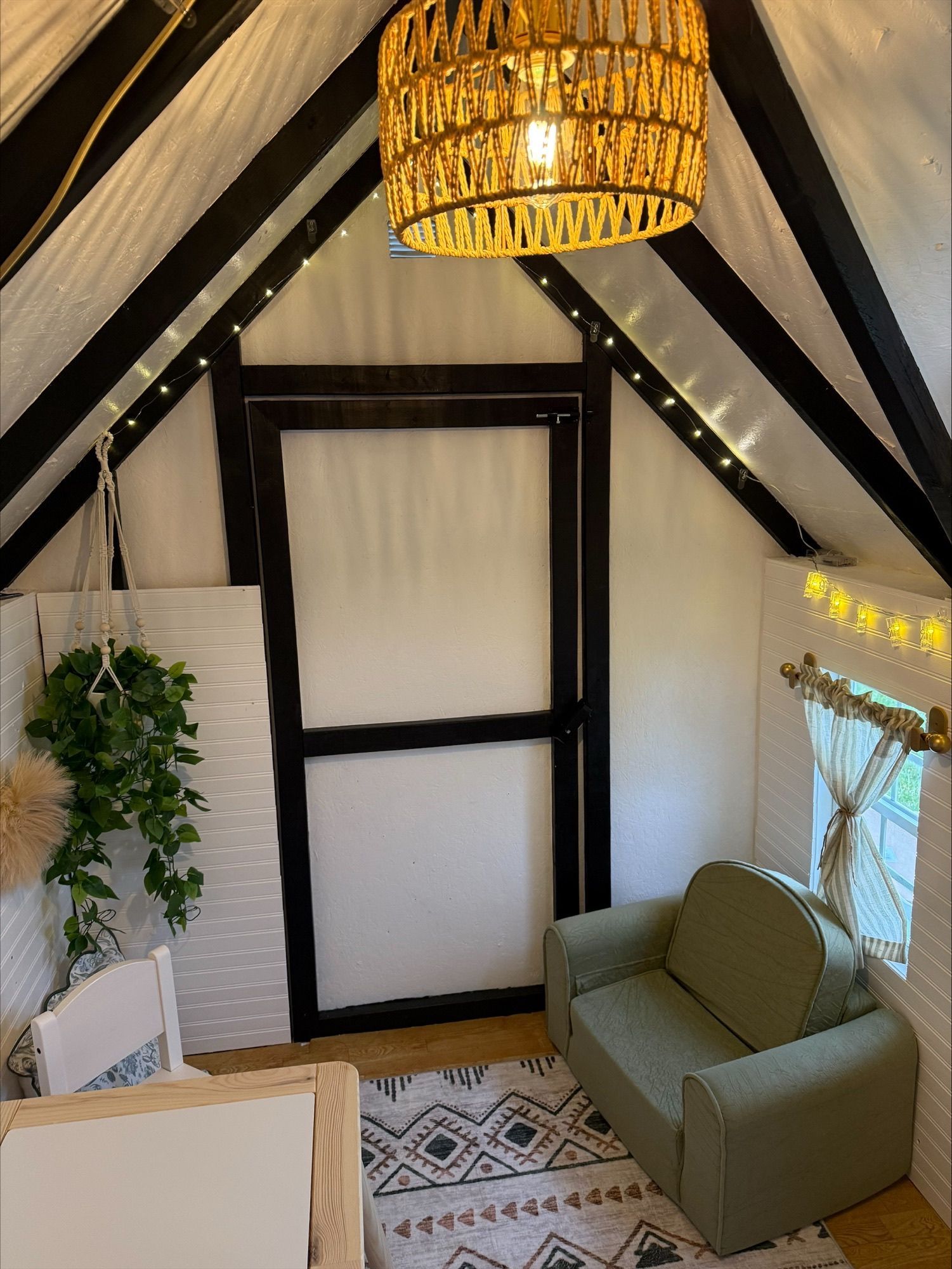 Cozy attic nook with black beams, wicker pendant light, sofa, plants, and patterned rug
