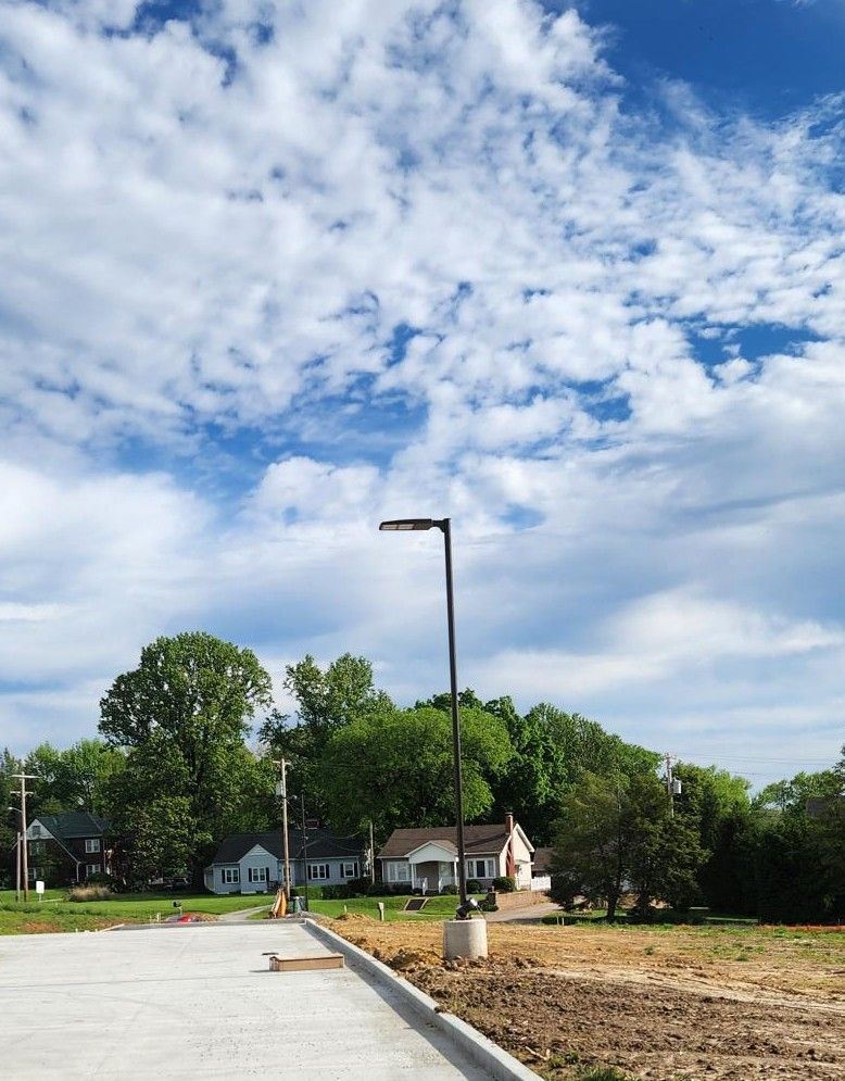 New sidewalk beside a dirt lot with a streetlamp, trees, and houses under a partly cloudy sky