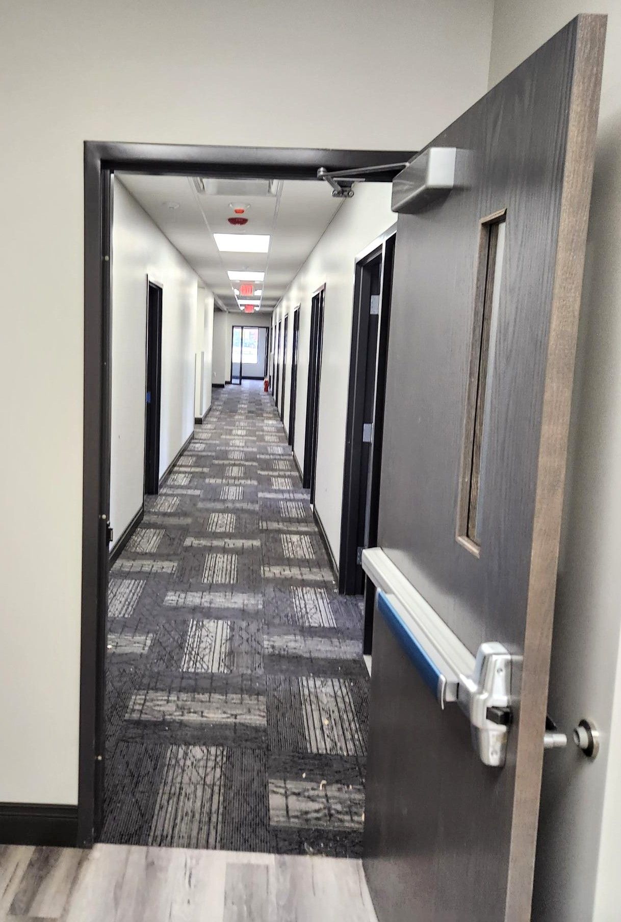 Open doorway to a long office hallway with patterned carpet and doors on both sides.