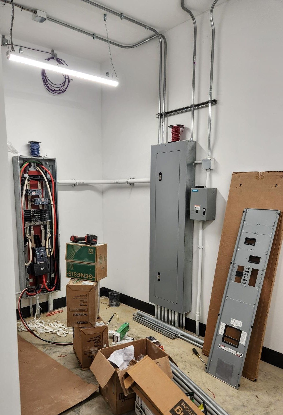 Electrical workshop with open panel, gray breaker box, conduit pipes, and scattered boxes on the floor.