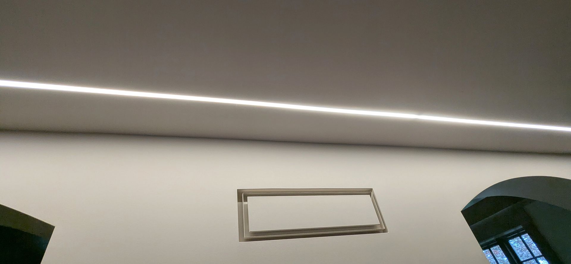 White wall with a rectangular frame and a ceiling light strip above, near a dark window on the right