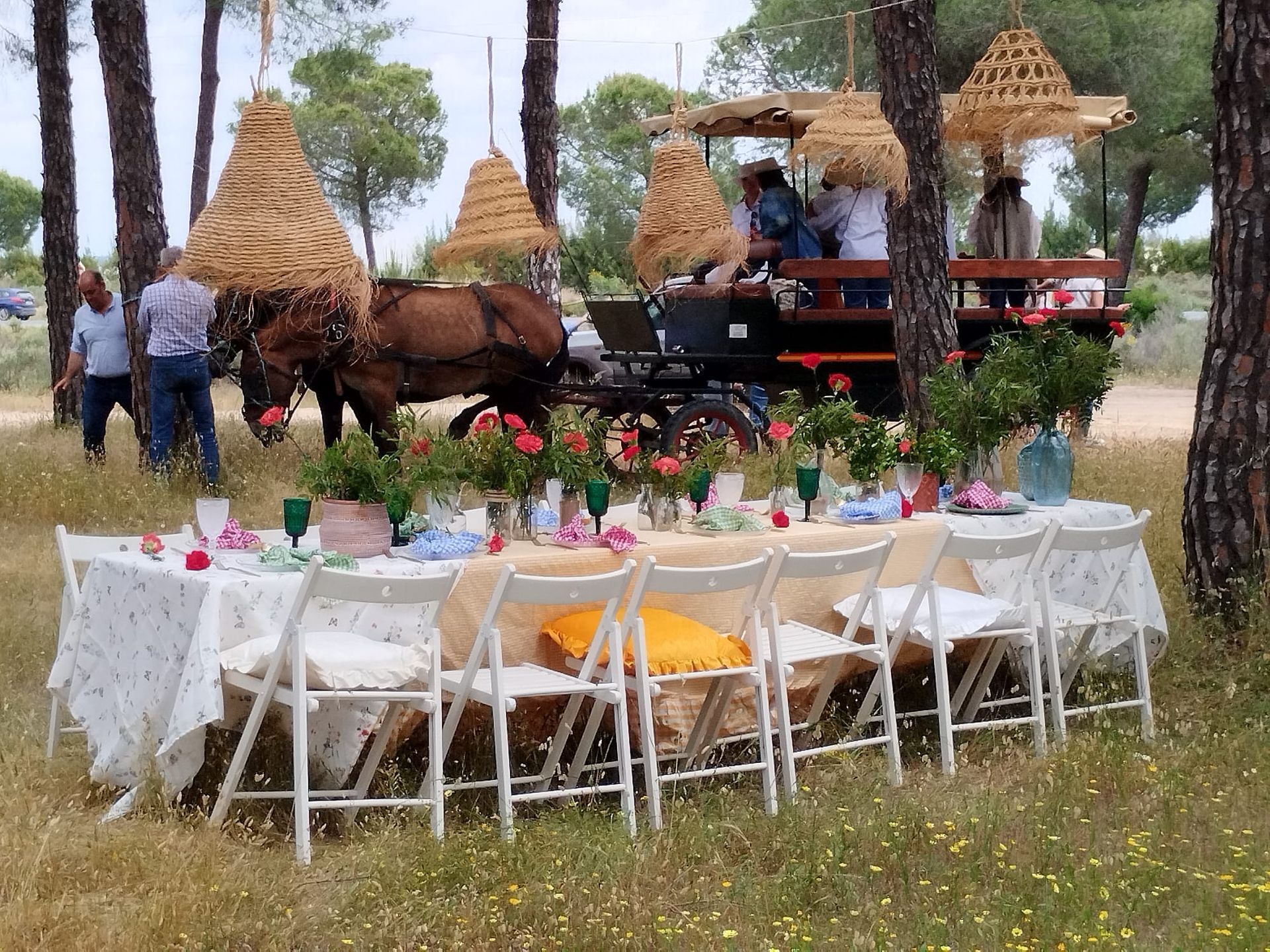 Un carruaje tirado por caballos y una mesa de comedor decorada en un campo. Los invitados están sentados a la mesa con flores, y los árboles rodean la escena.