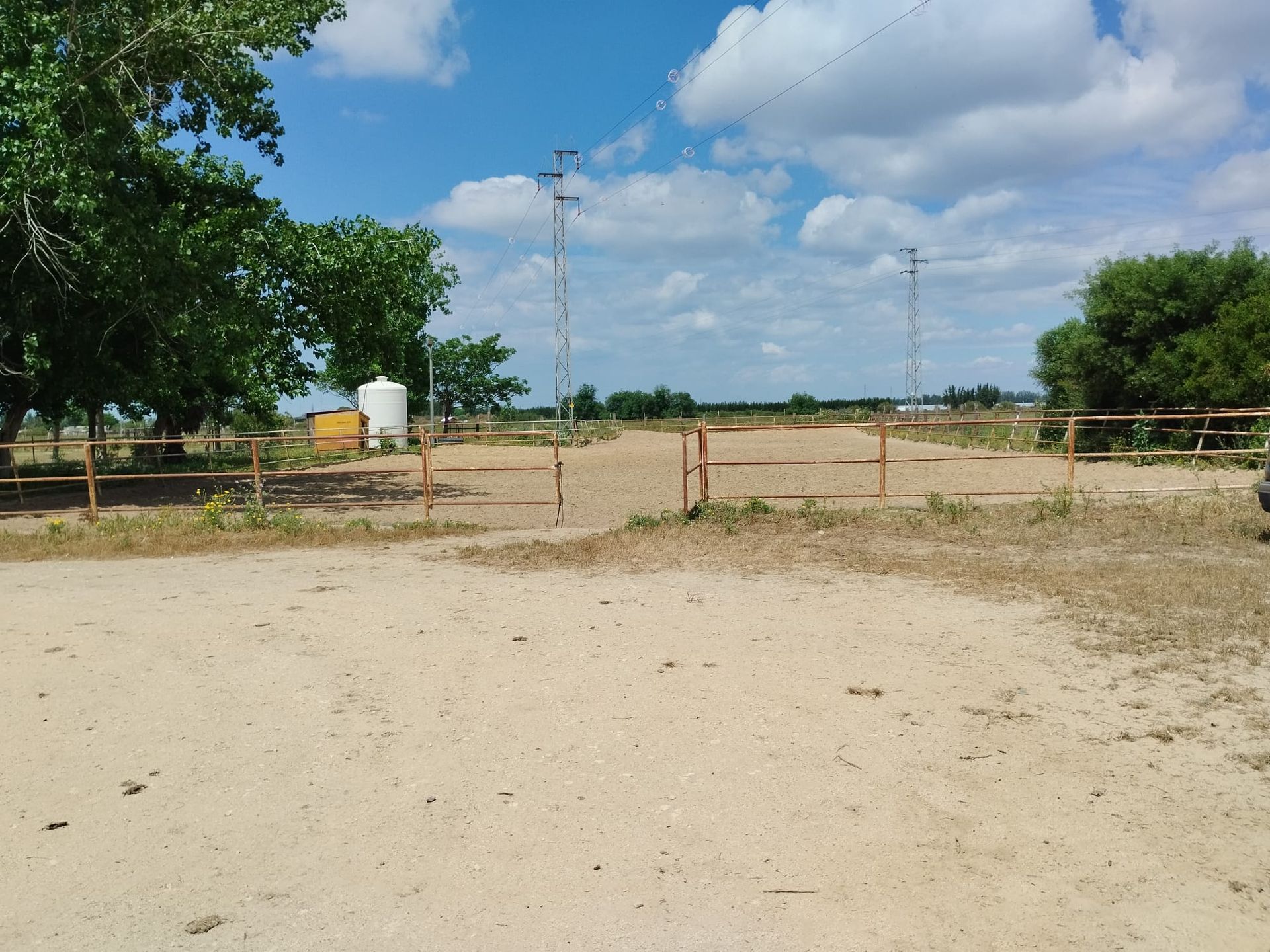 Portón abierto que conduce a un campo de arena con estructuras metálicas, bordeado de vallas de madera y árboles bajo un cielo parcialmente nublado.