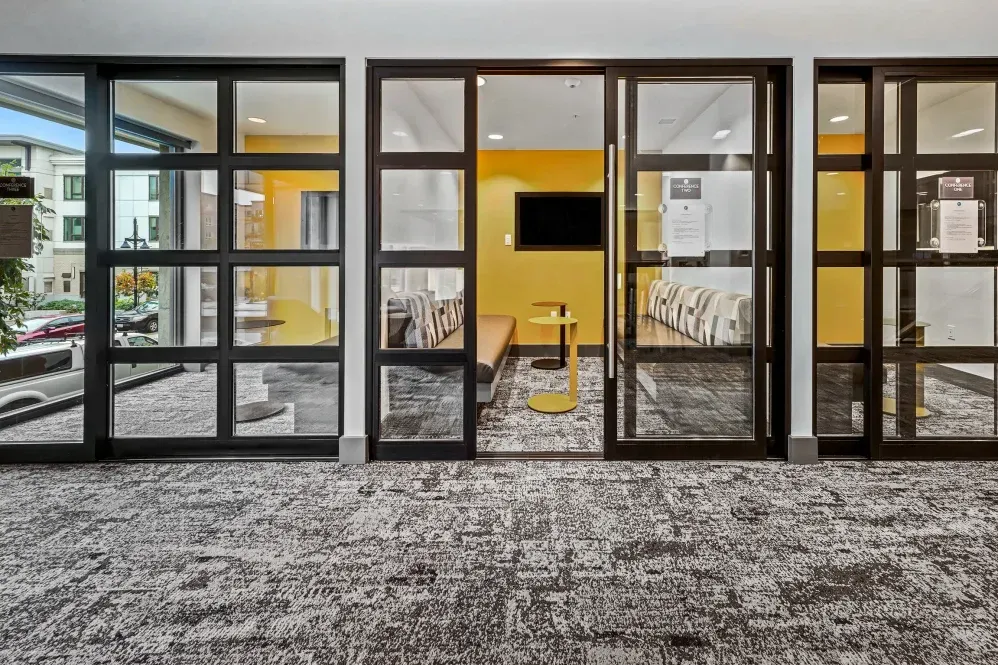 Glass doors open to a yellow-walled lounge area with seating, a TV, and a side table at The Verge in Auburn, WA.