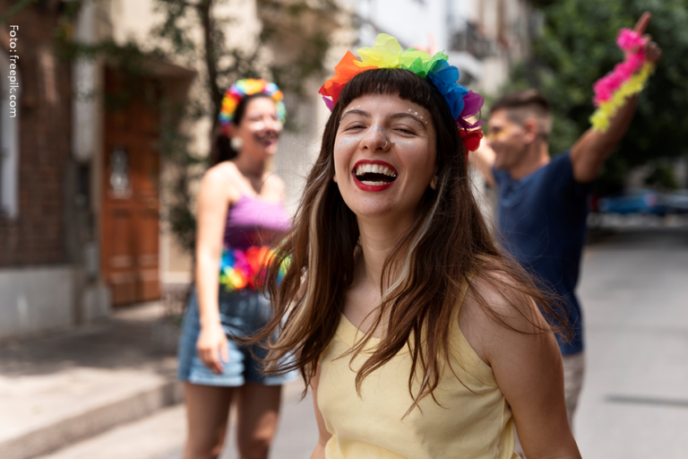 Mulher com coroa de arco-íris ri, outras pessoas com camisetas tie-dye comemoram na rua.