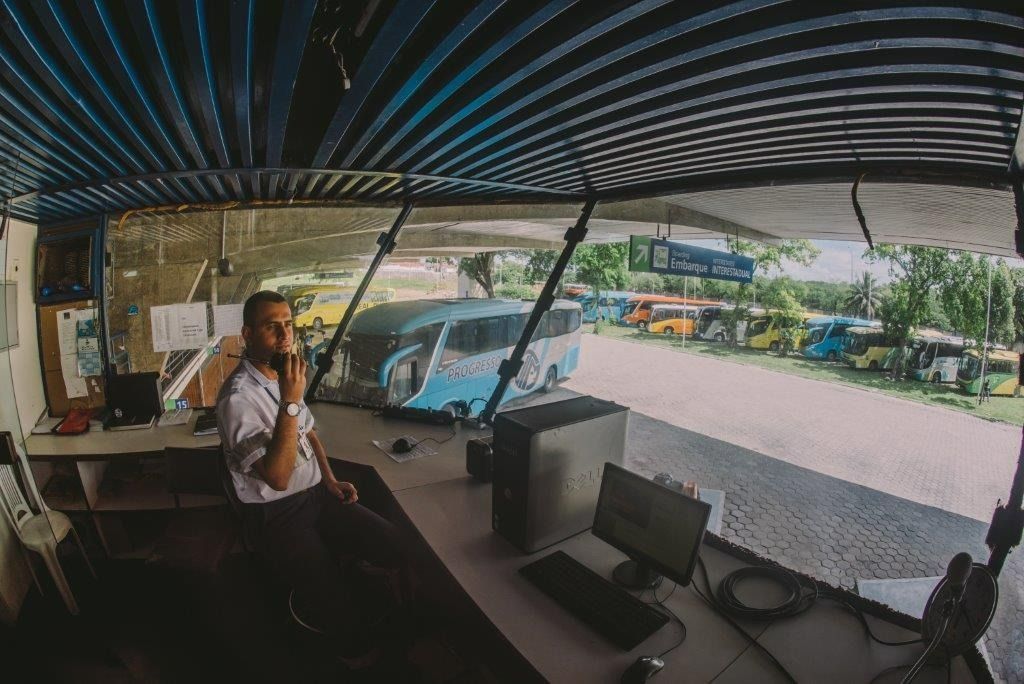Homem na sala de controle falando ao telefone, supervisionando ônibus estacionados ao ar livre sob abrigo.