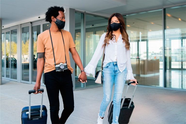 Casal com malas e máscaras sai de um prédio de mãos dadas. Dia ensolarado.