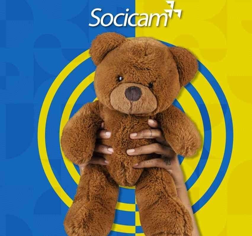 Mãos segurando um ursinho de pelúcia marrom contra um fundo azul e amarelo com o logotipo da Socicam