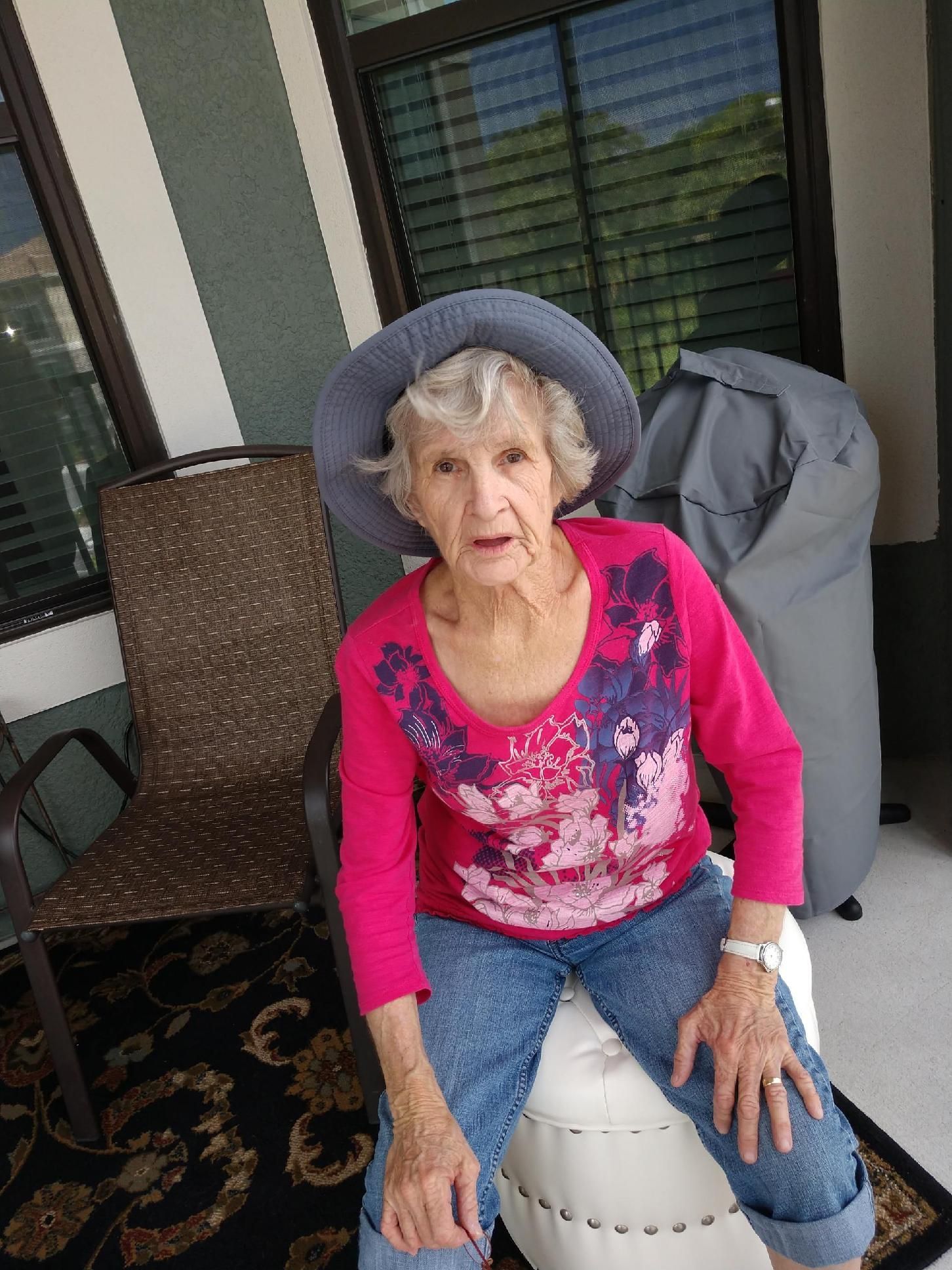 An elderly woman wearing a hat and a pink shirt is sitting on a chair on a porch.