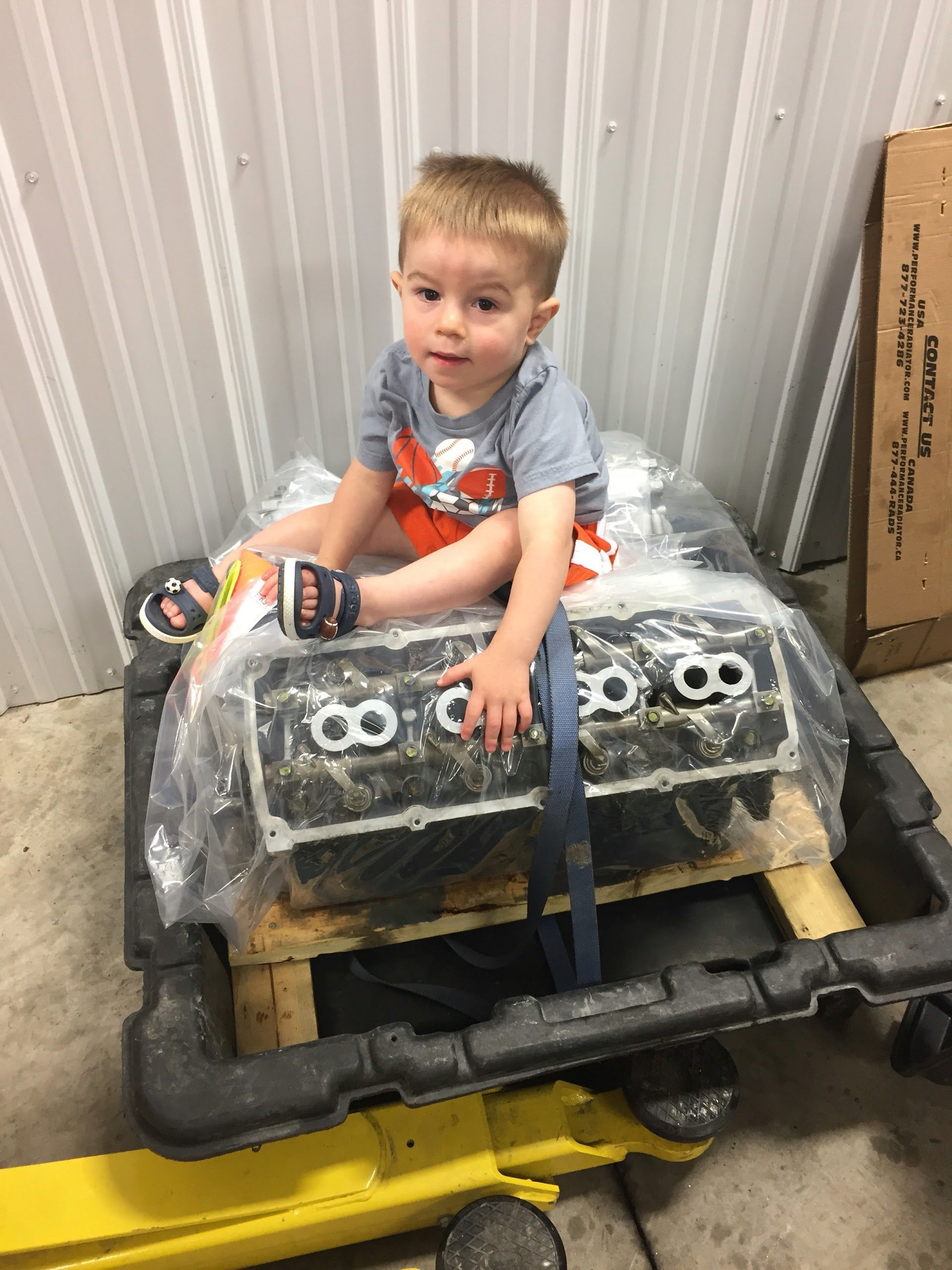 Child sitting on top of an engine wrapped in plastic, inside a garage | Central Auto Works