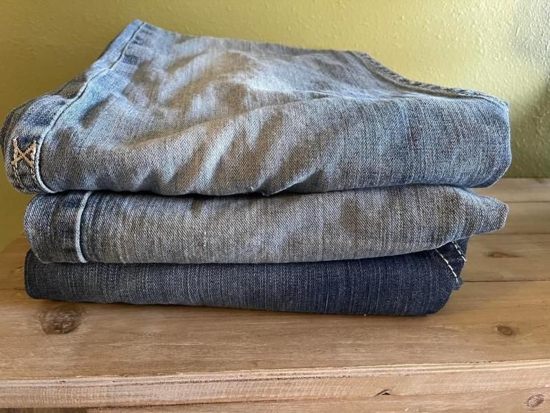 Stack of three folded denim jeans on a wooden surface; varying shades of blue.