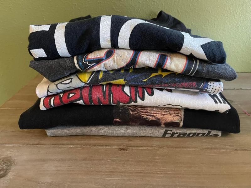 Pile of folded t-shirts, various colors and graphic designs, on a wooden surface.