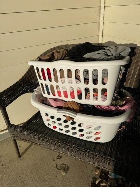 Two white laundry baskets filled with clothes stacked on a wicker chair.