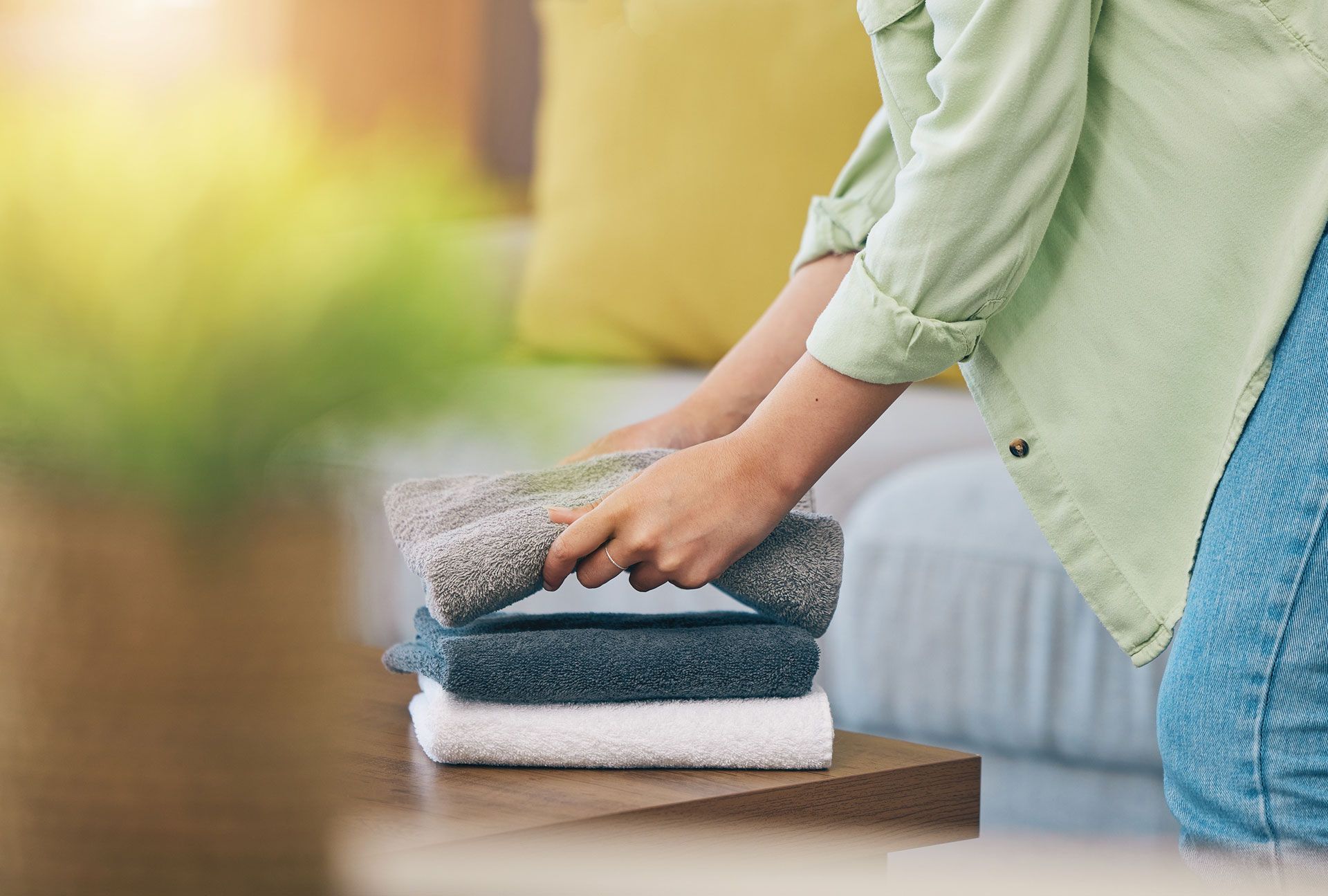 Person folding towels on a wooden table; blurred background with couch and sunlight.