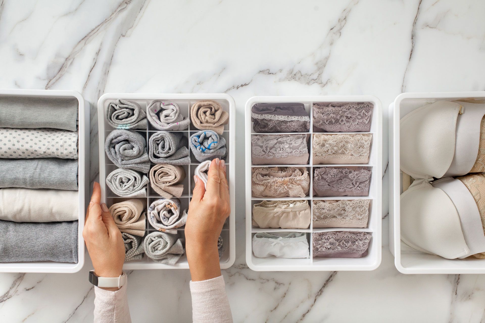 Person organizing folded clothing in white storage bins on a marble surface.