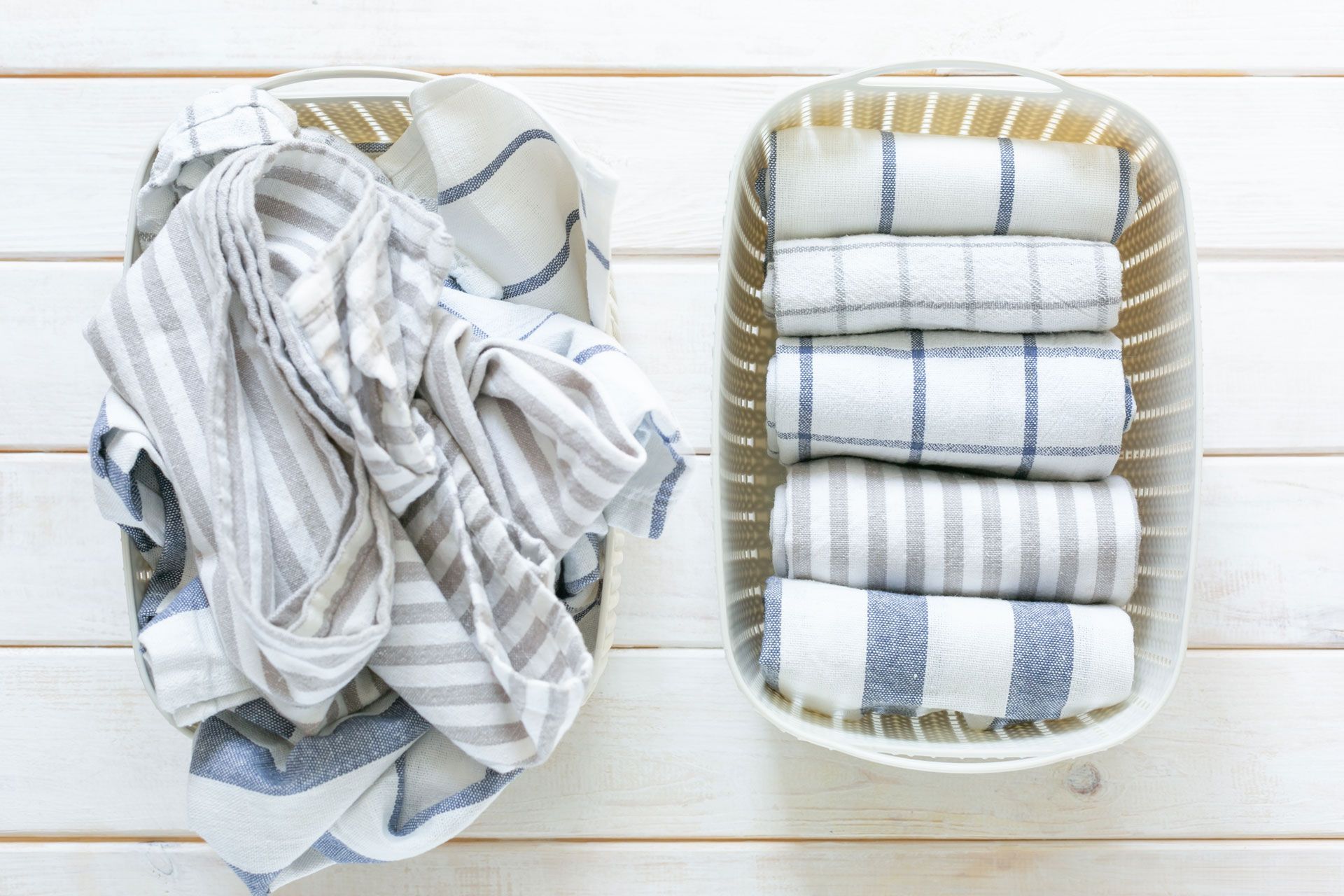 Two white baskets on a wooden surface; one holds crumpled striped towels, the other holds neatly rolled towels.